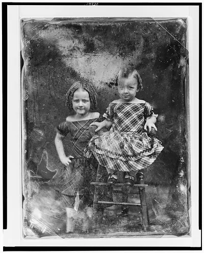 [Two unidentified girls, about three and five years of age, the younger child seated in high chair, older one standing beside her on left, both nearly facing front]