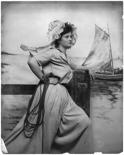 [Woman wearing bonnet, holding rope and standing on pier; fishing boat in background]