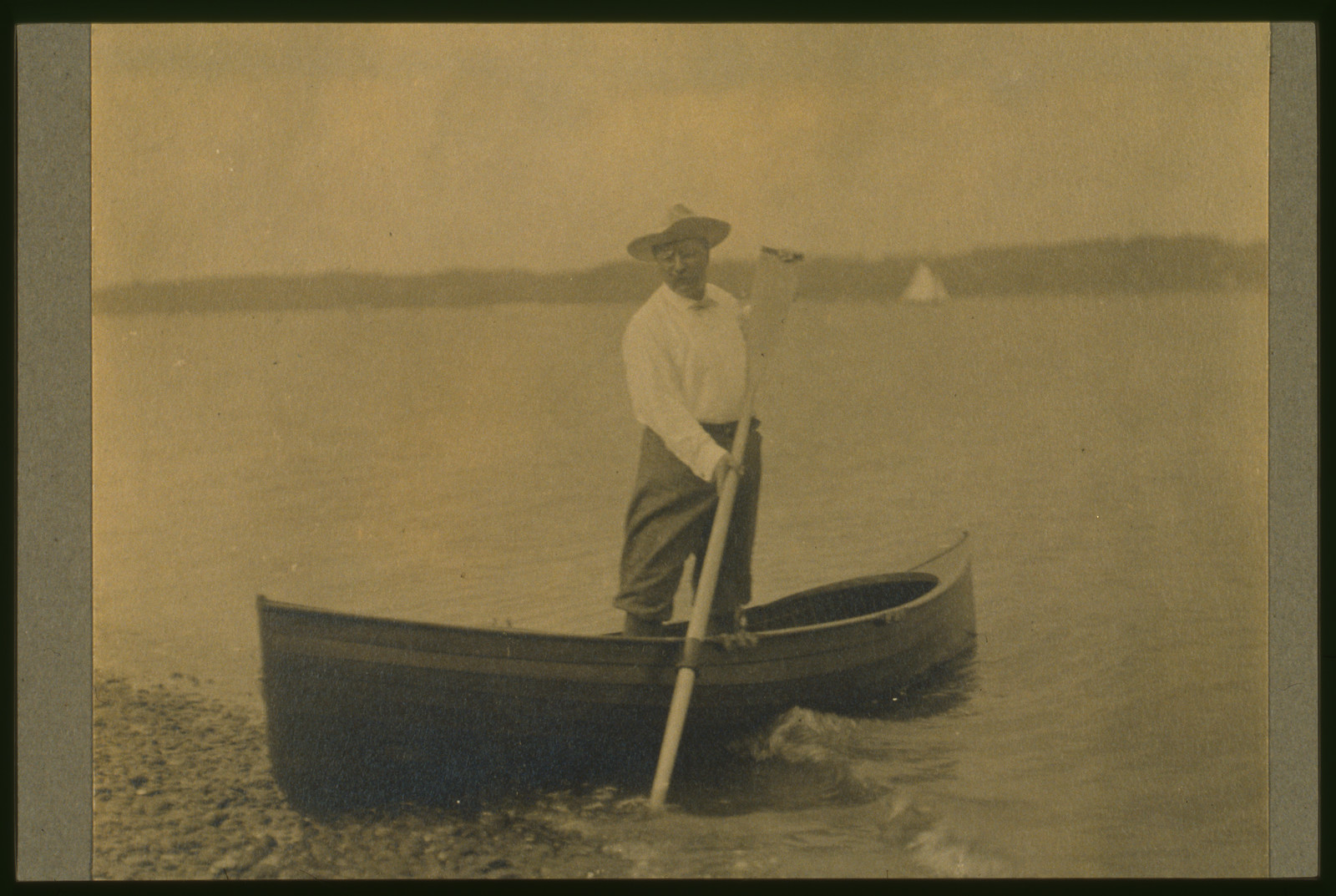[Theodore Roosevelt, standing up in rowboat, pushing away from shore with oar], c1905. — historic photograph