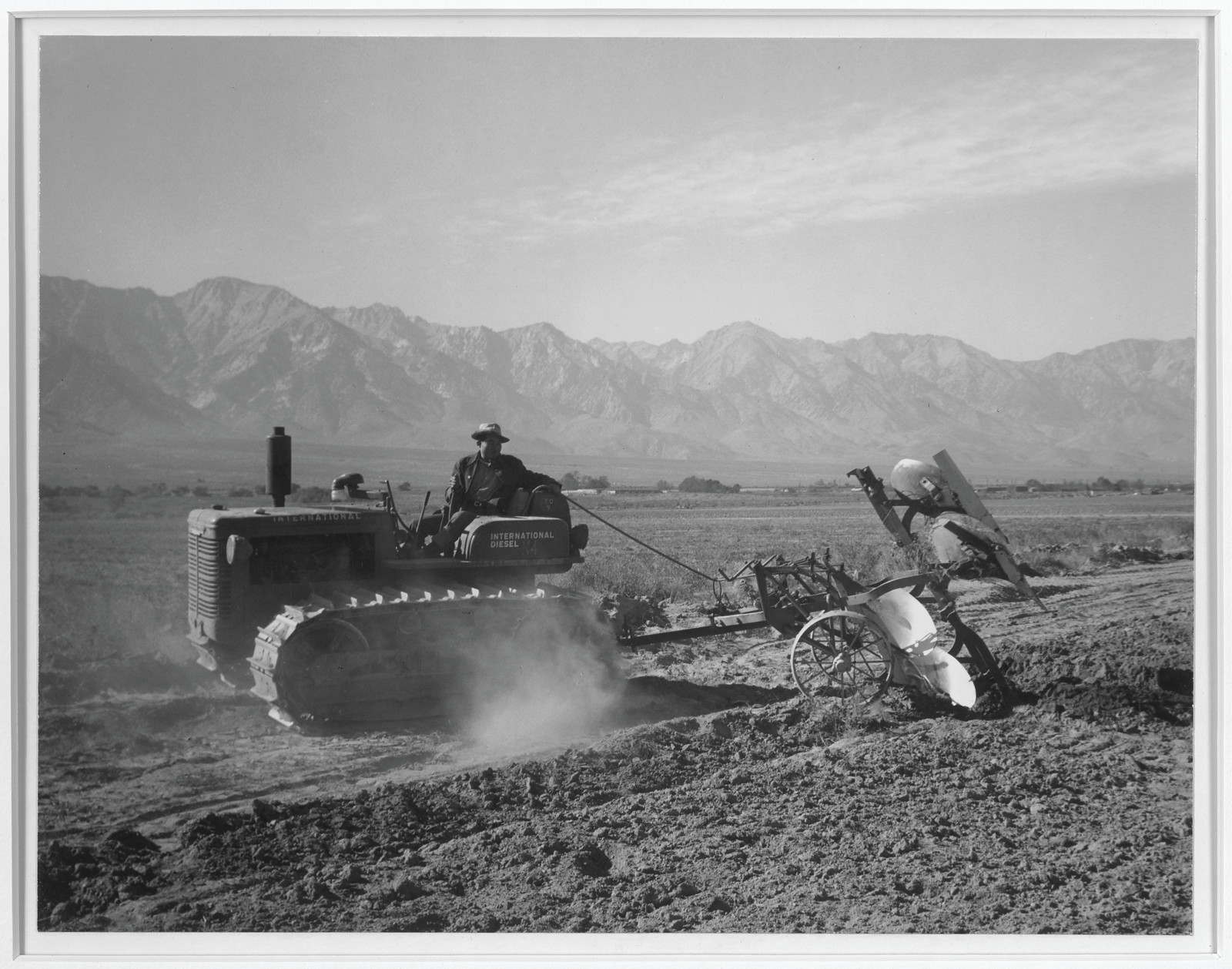 Crawler tractor pulling implement across field, mountains behind — historic image
