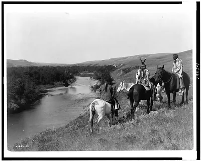 [Three Piegan Indians and four horses on hill above river]