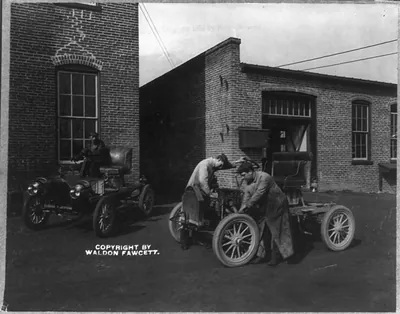 [2 men working on an automobile outside of automobile plant]