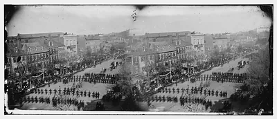 Military column marching down a city avenue, crowds on sidewalks
