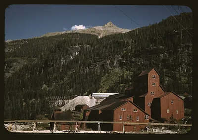 Red mill buildings and tailings pile below forested mountain slopes