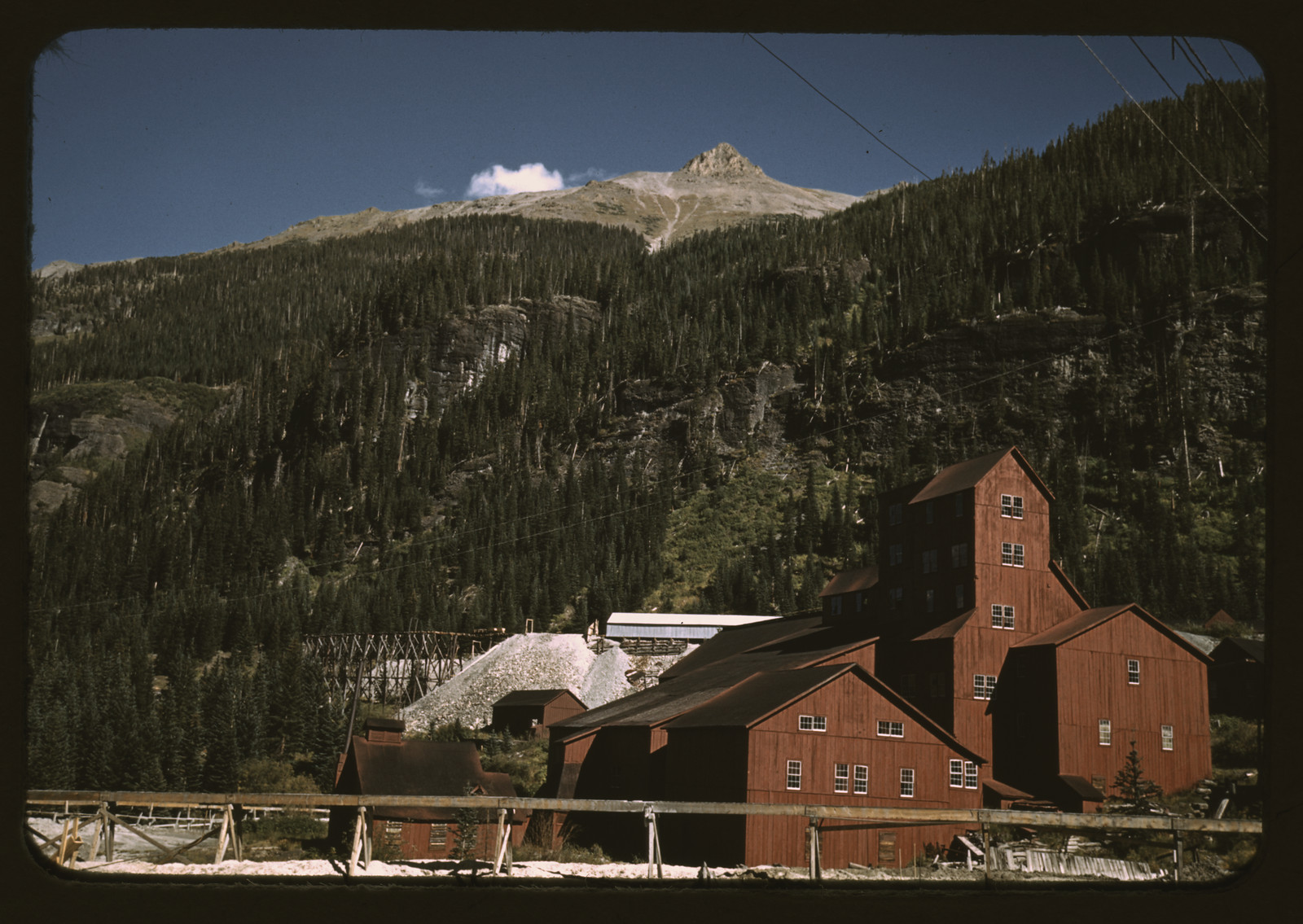 Red mill buildings and tailings pile below forested mountain slopes — historic image