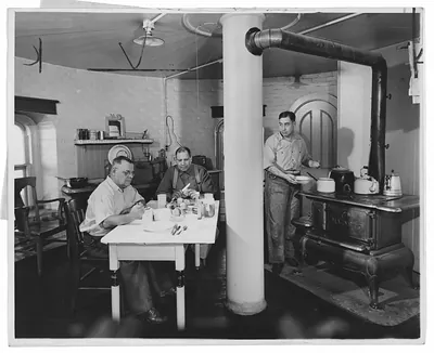 Two men eating at a table beside a woodstove, with a standing man
