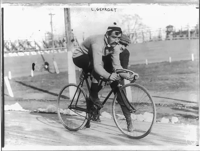 [Leon Georget, full length, seated on bicycle, facing right; another man holding the bicycle]