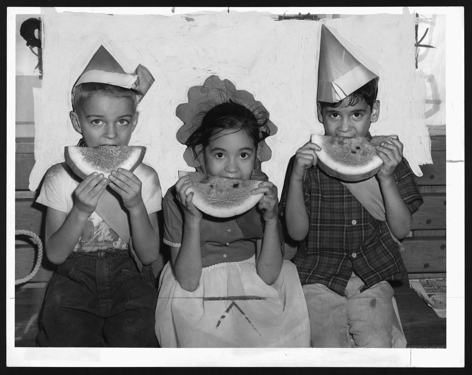 Three children eating watermelon, wearing paper hats — historic image