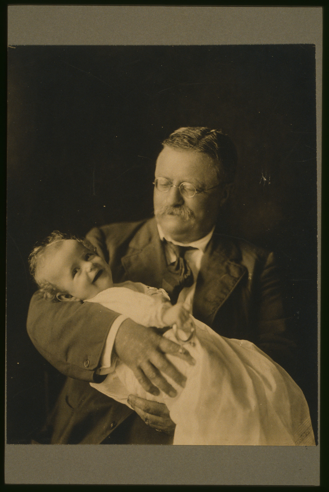 [Theodore Roosevelt, half-length portrait, seated, looking down, holding his grandchild, Kermit Roosevelt, Jr.], c1916. — historic photograph