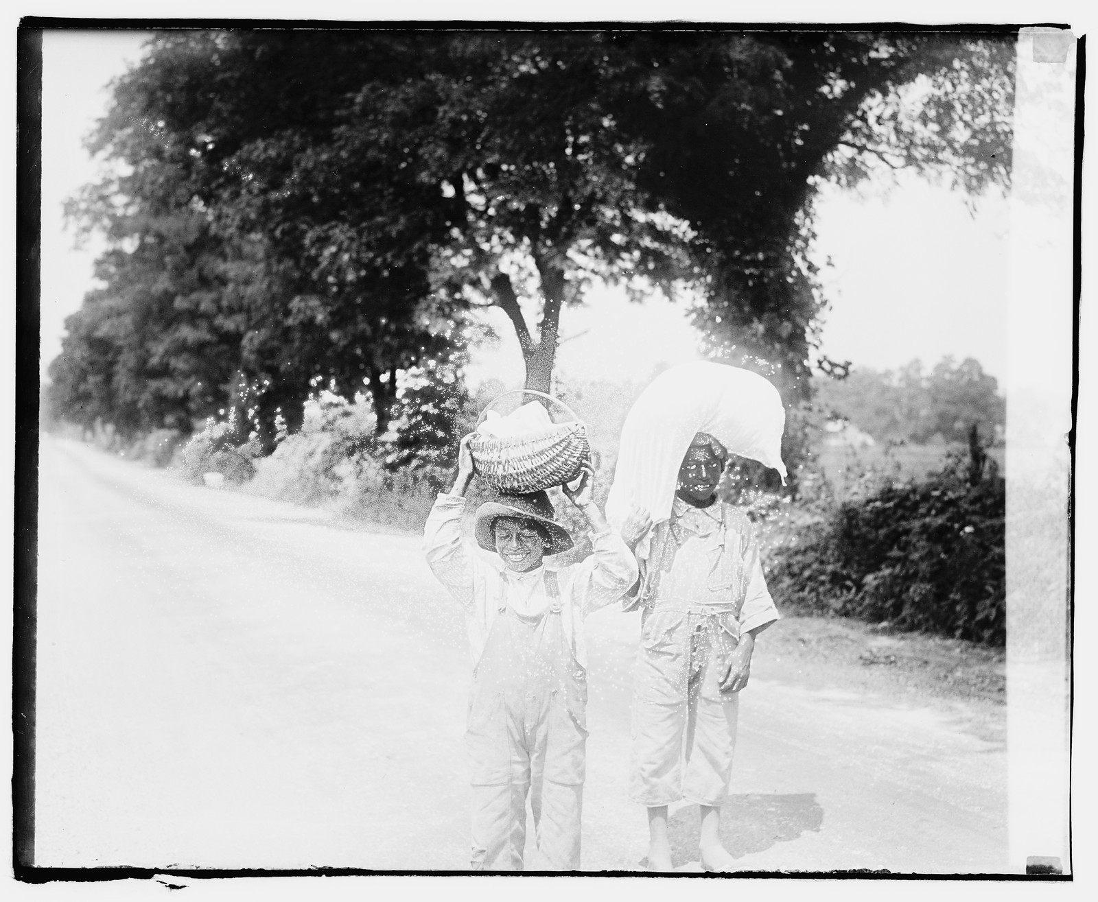[Two little boys in overalls standing in a dirt road, balancing loads on their heads], [1920] — historic photograph