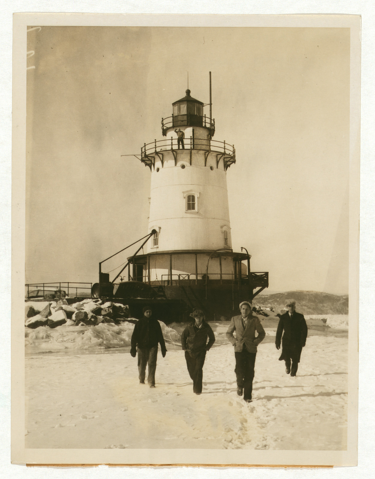 Four men walking on snow-covered ice by a lighthouse — historic image