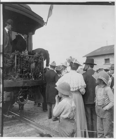 [Theodore Roosevelt, leaning over to shake hands with person from rear platform of railroad car]
