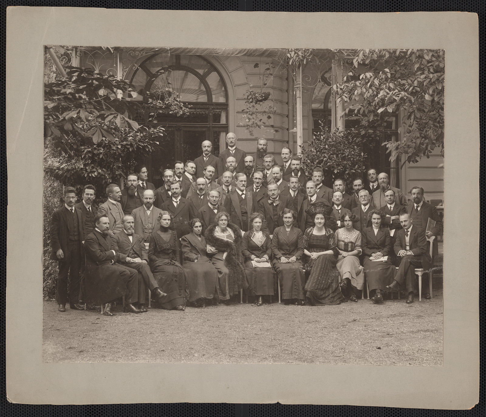 Large group photograph of men and women seated and standing outdoors before a building — historic photograph
