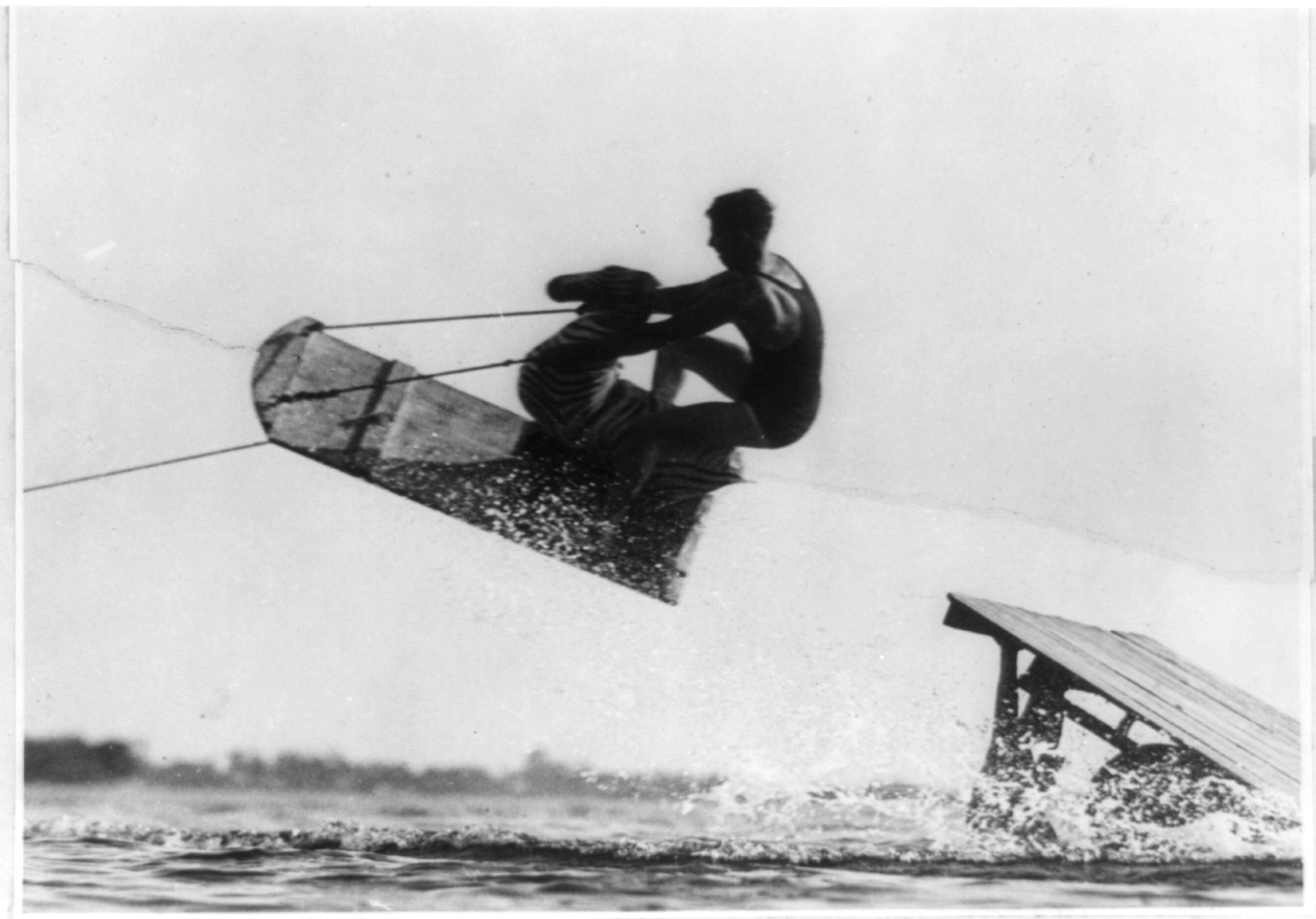 Man aquaplaning on a wooden board, jumping a ramp — historic image