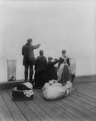 [Four immigrants and their belongings, on a dock, looking out over the water; view from behind]