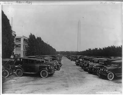Two rows of automobiles parked at the rear of the Navy Bldg. In the distance are the Washington Monument and the dome of the Capitol
