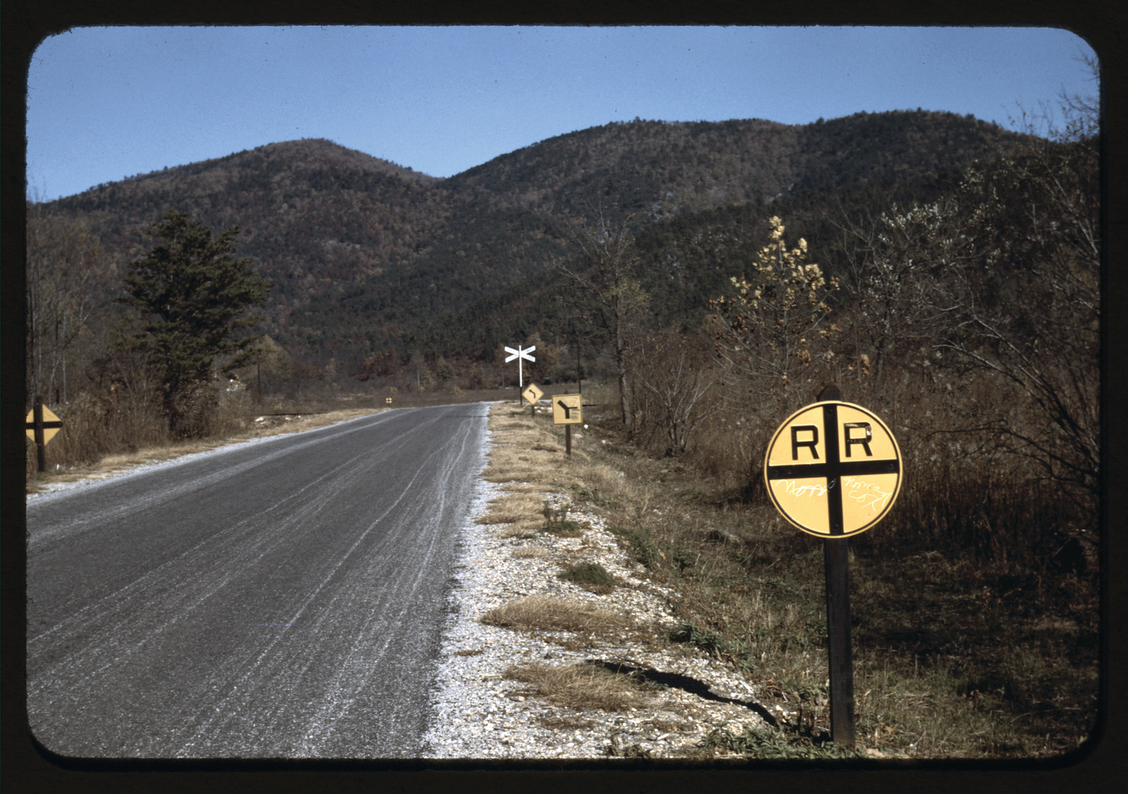 Road with railroad crossing signs below wooded mountains — historic image