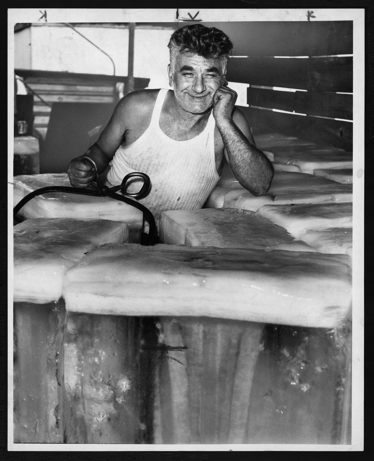 Smiling ice handler with tongs leaning on ice blocks — historic image
