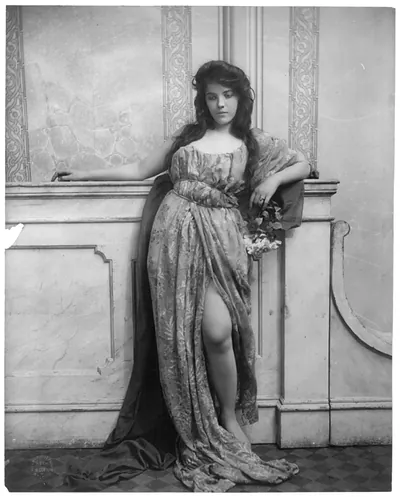 [Young woman modeling: full lgth., standing, leaning on mantle and waring split skirt]