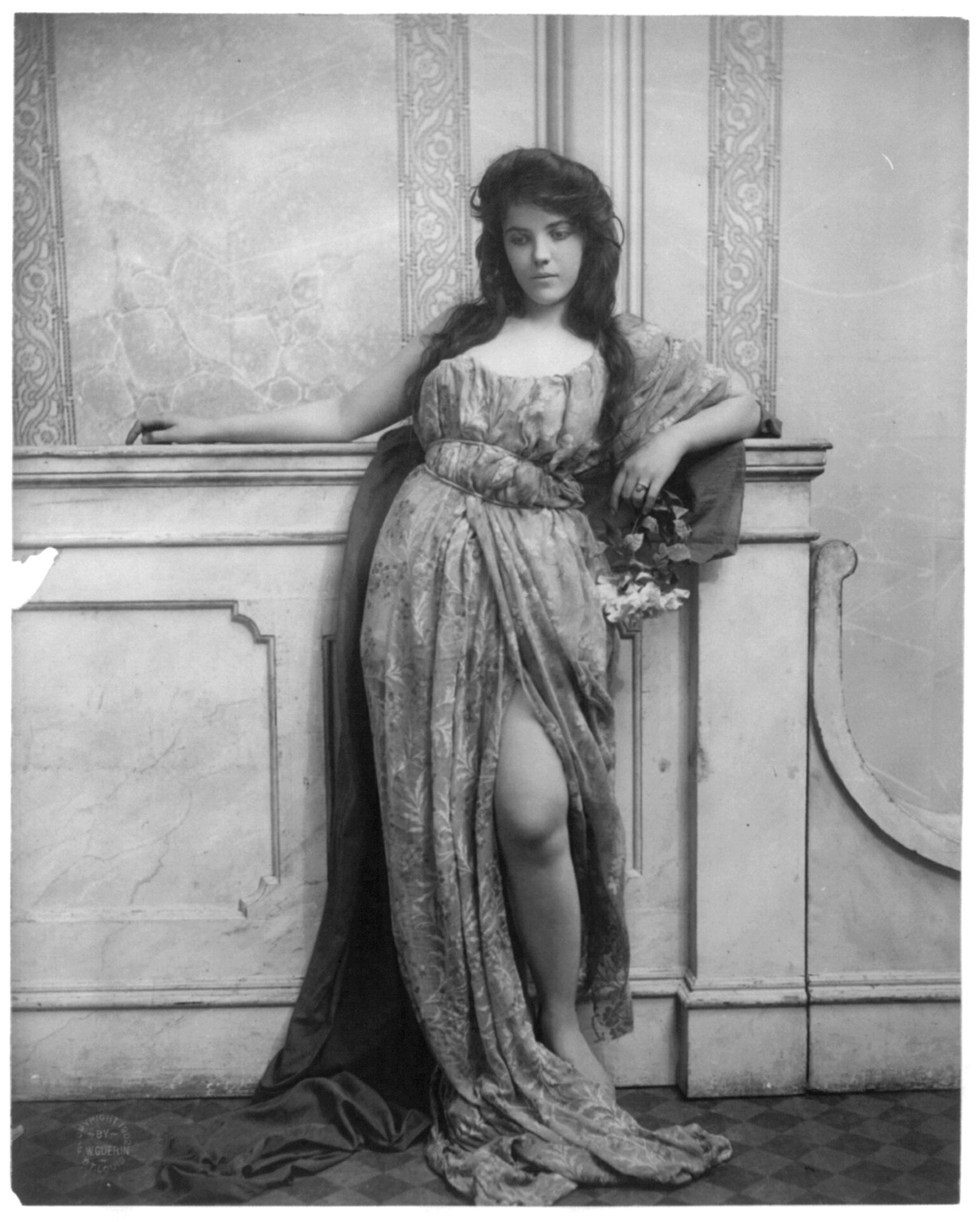[Young woman modeling: full lgth., standing, leaning on mantle and waring split skirt], c1902. — historic photograph