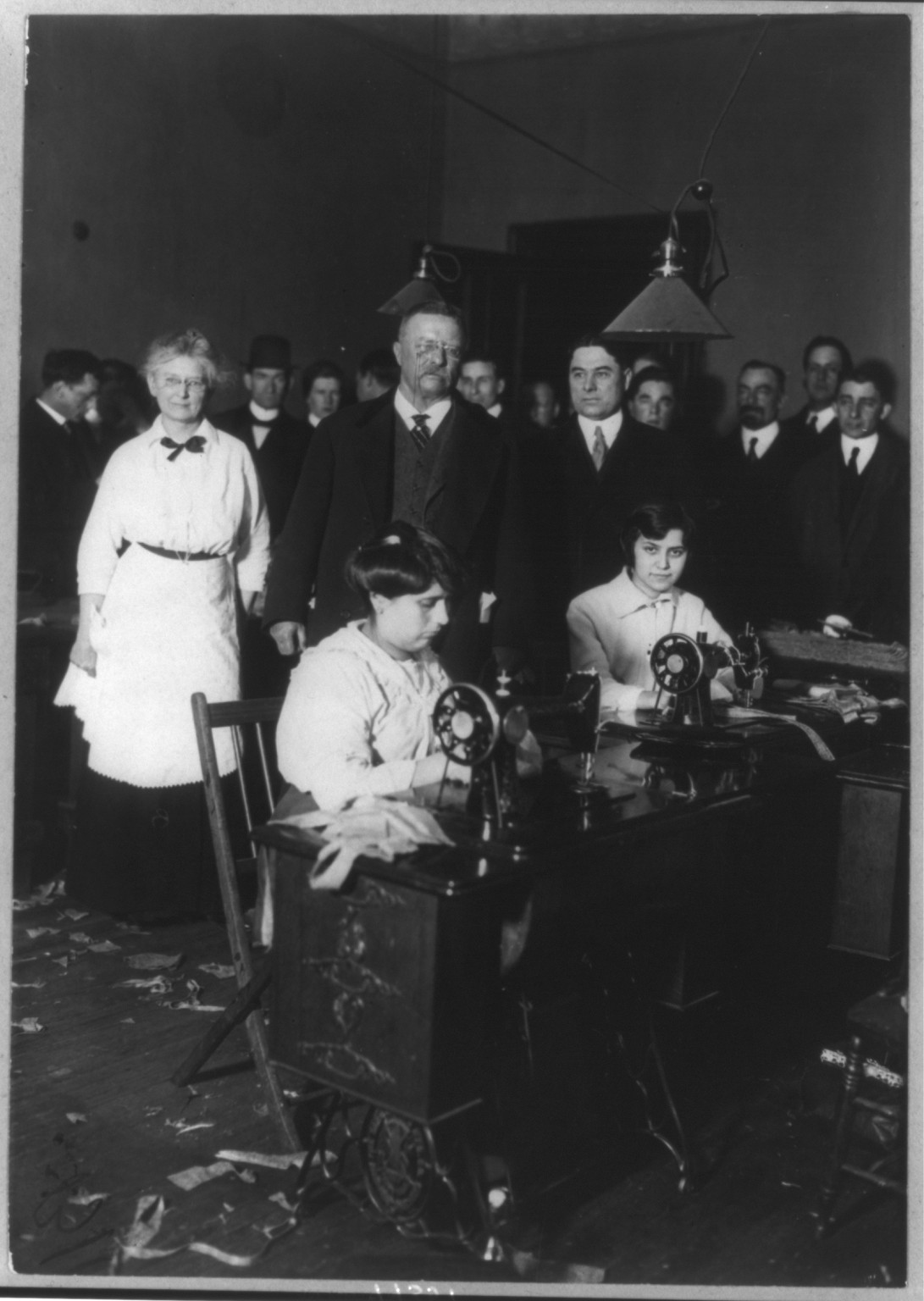 Women operating sewing machines in a workroom with standing observers — historic image