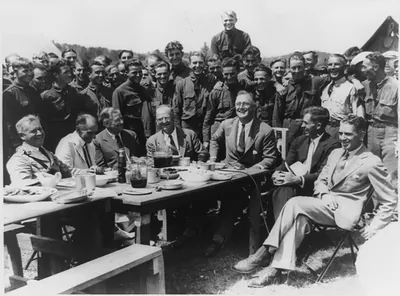 [President Franklin D. Roosevelt having lunch at mess table in Camp Fechner, a Civilian Conservation Corps camp, at Big Meadows, Va., with General Paul B. Malone; Col. Louis Howe; Secretary Ickes of the Interior Department; Robert Fechner, Director of the Civilian Conservation Corps; Secretary Wallace of Agriculture Dept.; and Assist. Secretary of Agriculture Tugwell]