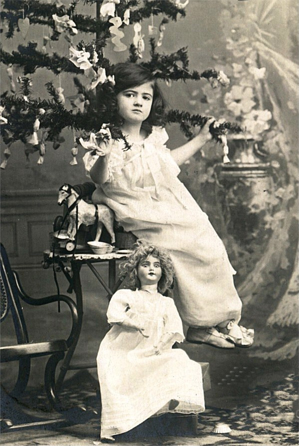Girl in white dress beside Christmas tree and doll — historic image