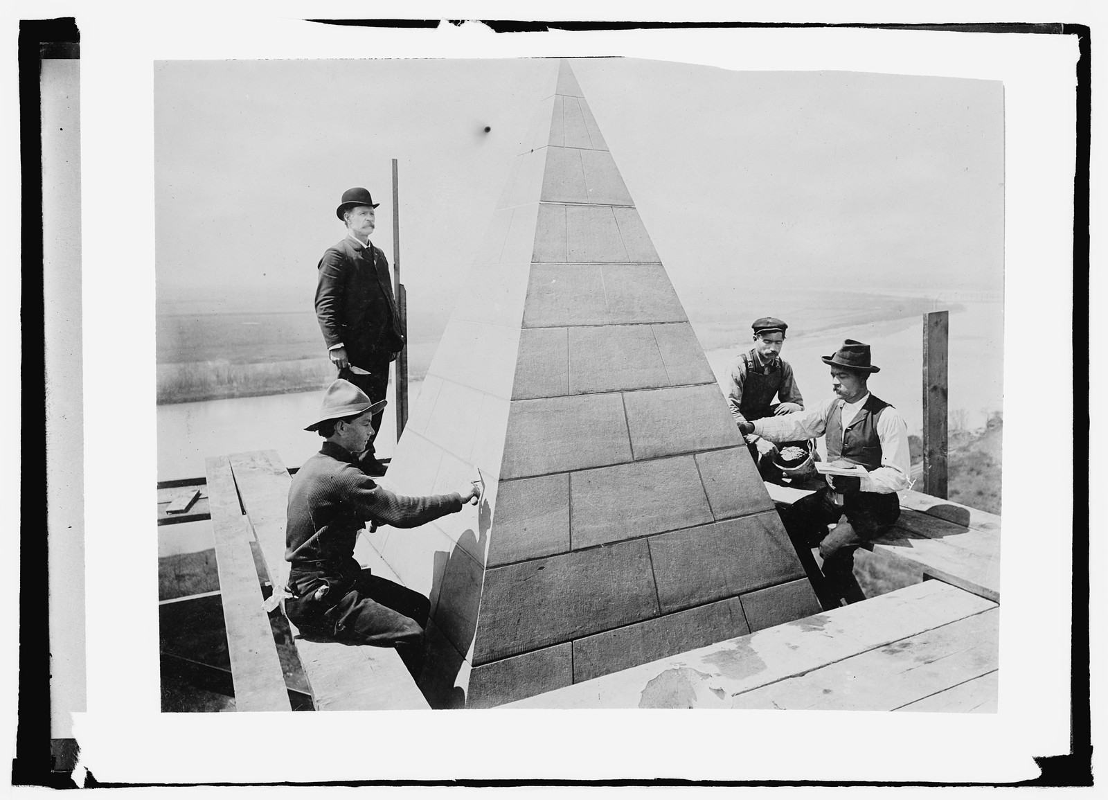 Men working on pyramidal monument capstone above river — historic image