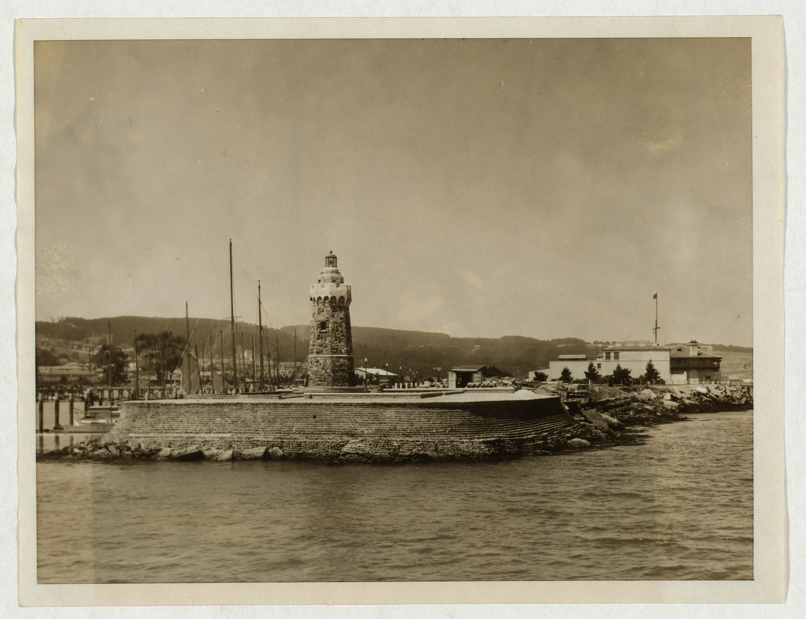 Stone lighthouse on a breakwater with sailboat masts — historic image