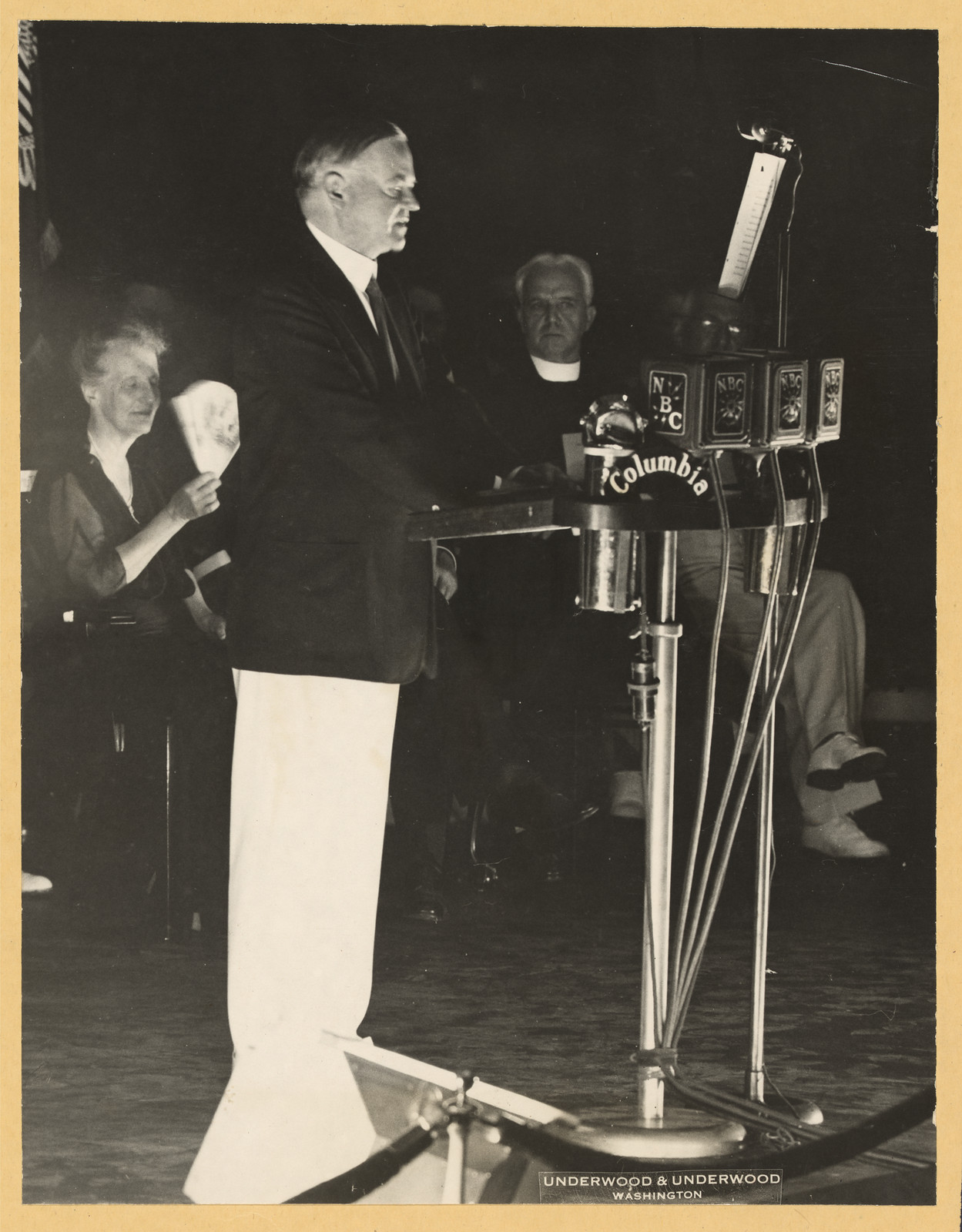 Speaker at NBC and Columbia radio microphones, with seated audience — historic image