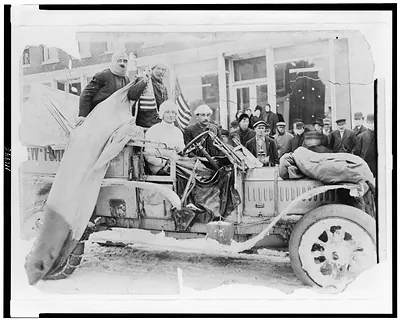 [Four men in automobile, with American flags on snow-covered street, on the New York to San Francisco leg of the 1908 New York to Paris automobile race]