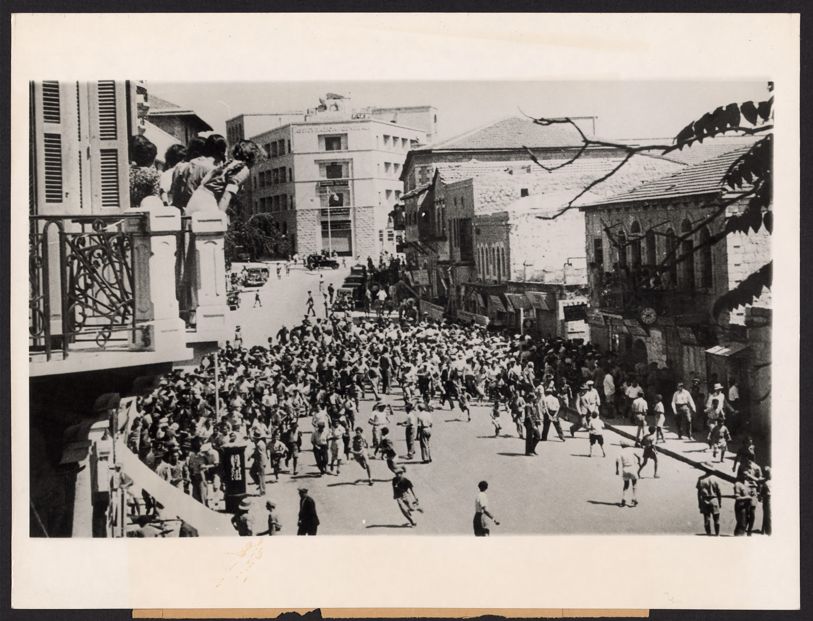 Crowd running through a city street, onlookers on balcony — historic image