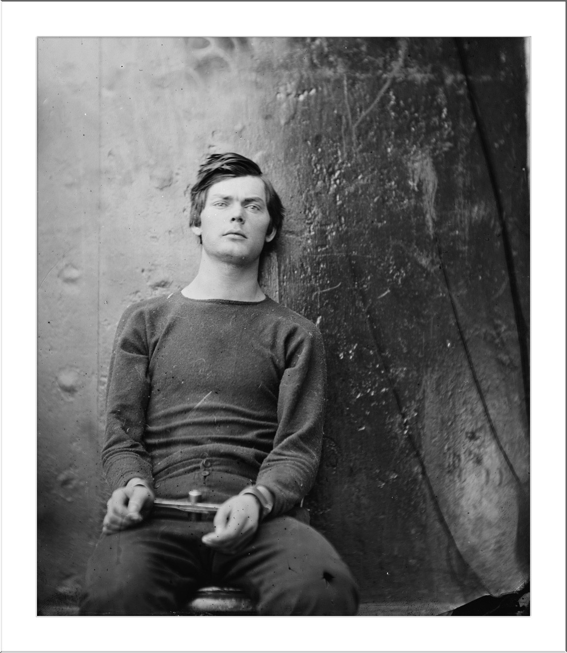 Seated young man in sweater against dark backdrop, hands clasped — historic image