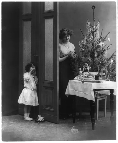 [Sentimental Christmas scenes of children and trees: small girl hiding behind door; woman is arranging toys around small tree on table]
