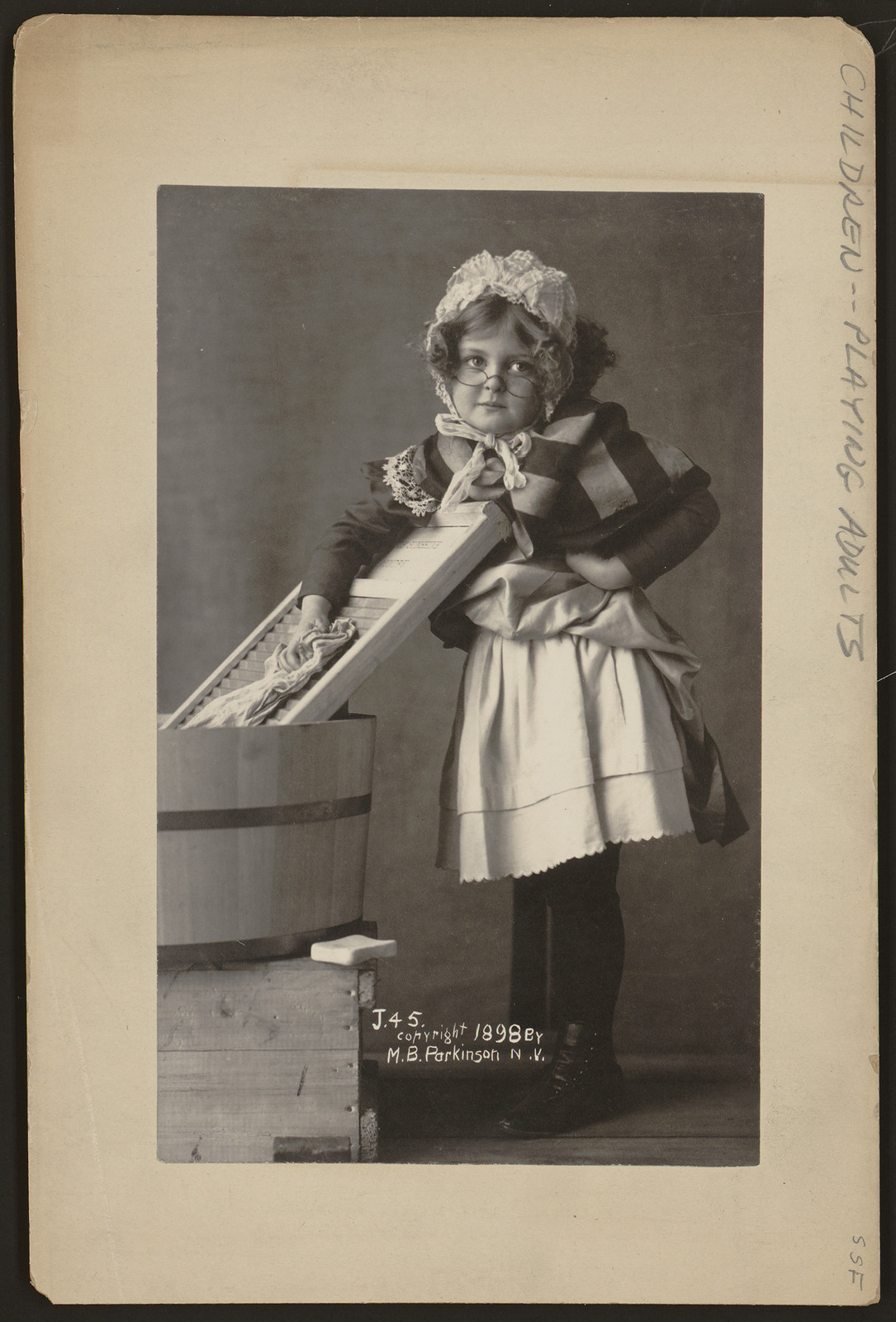 [Girl wearing eyeglasses, bonnet and apron, with washboard and tub], c1898. — historic photograph