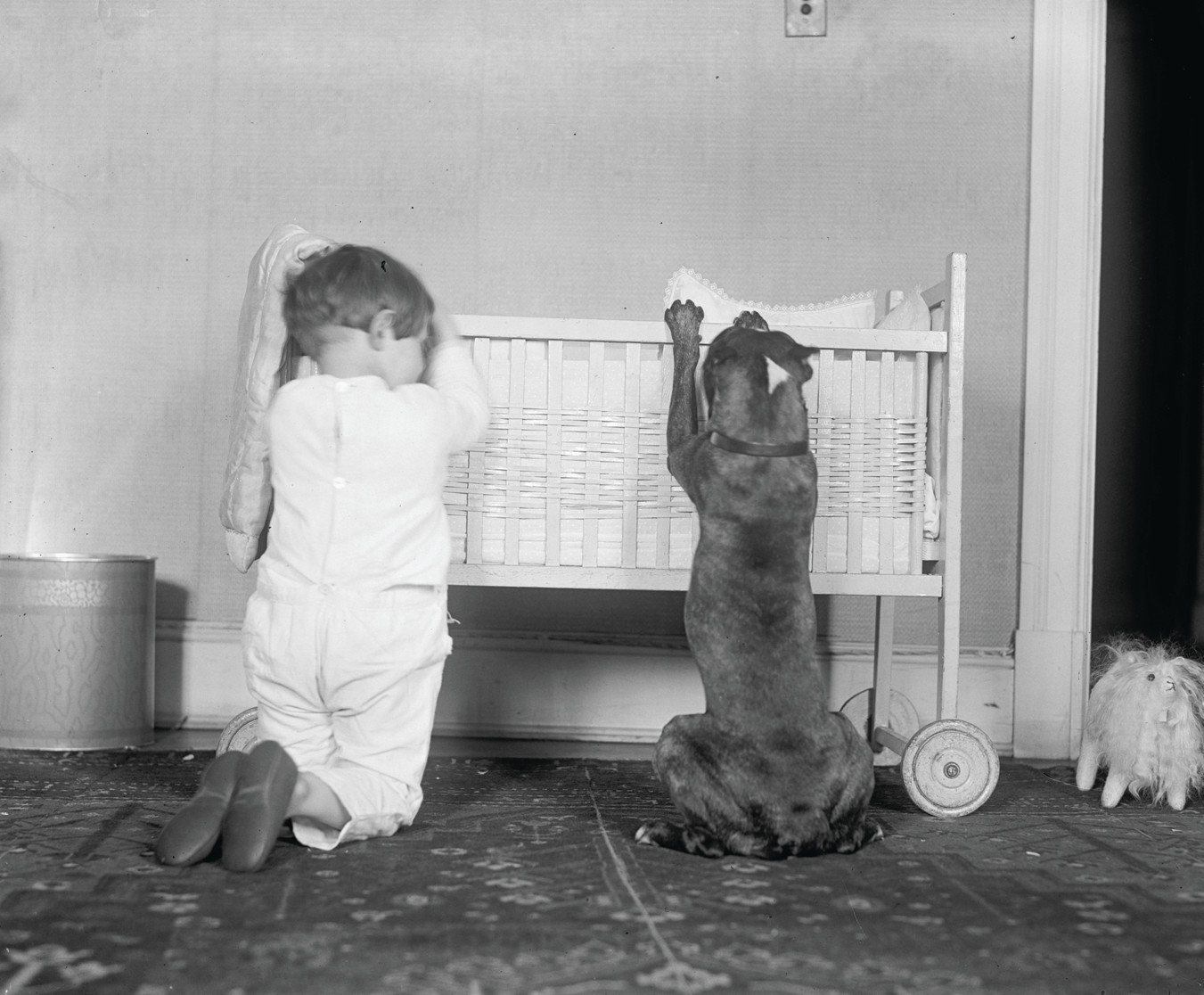 Toddler and dog beside a wicker crib — historic image