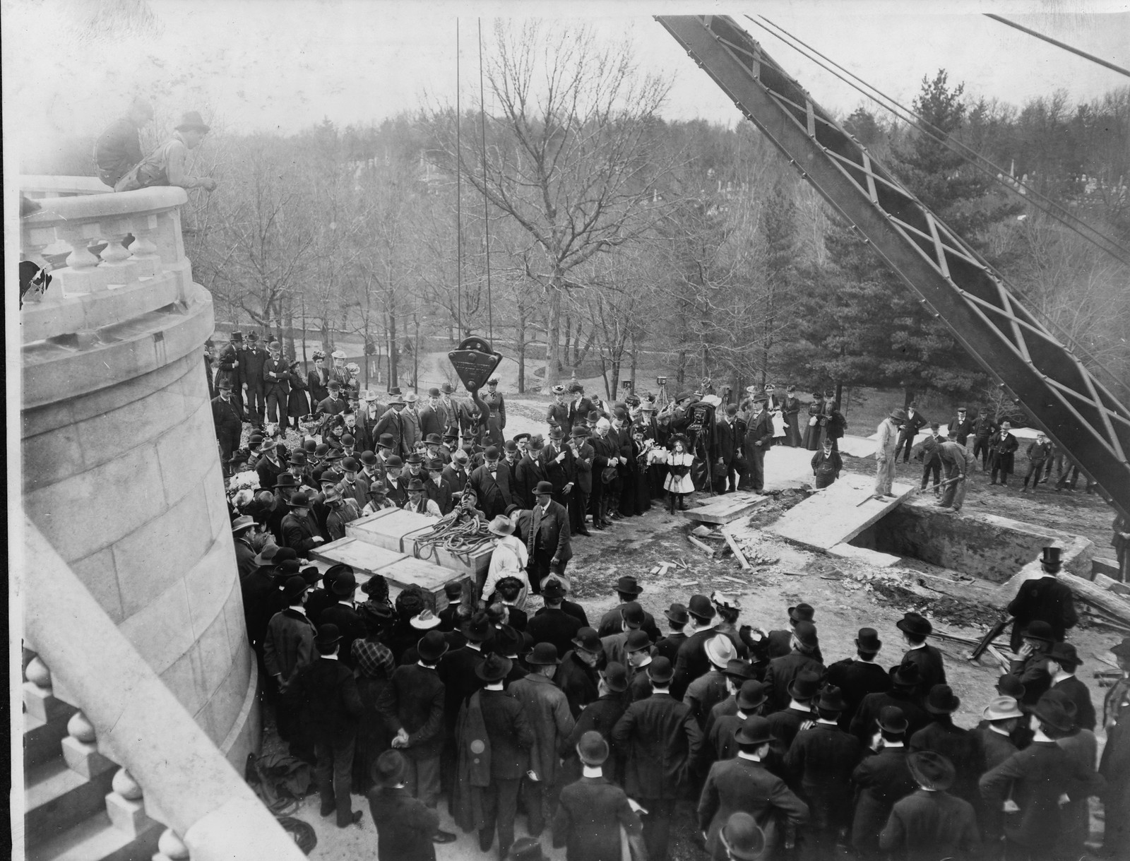 Crowd watching crane lift casket beside stone vault excavation — historic image