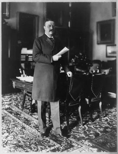 [Theodore Roosevelt, full-length portrait, standing by table, facing right; holding paper in right hand, left hand on back of chair]