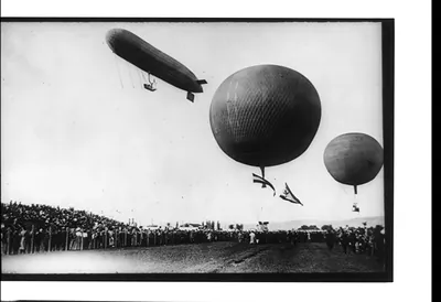 Dirigible and 2 balloons aloft above crowd