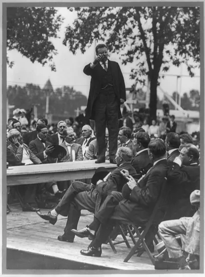 [Theodore Roosevelt standing on table outdoors and making speech to men seated around him, right hand raised] / Smith Art Photography, Freeport, Ill.