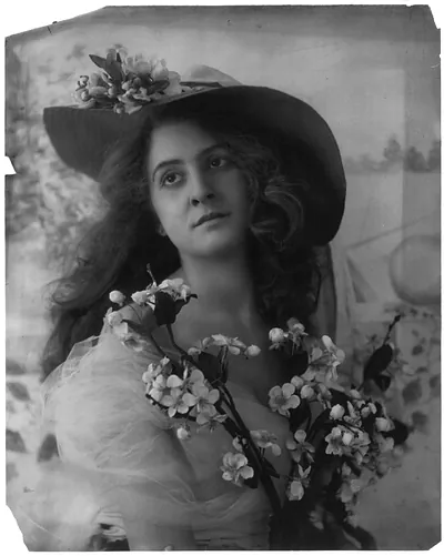 [Young woman modeling: half lgth., wearing large-brimmed hat and holding fruit tree blossoms]