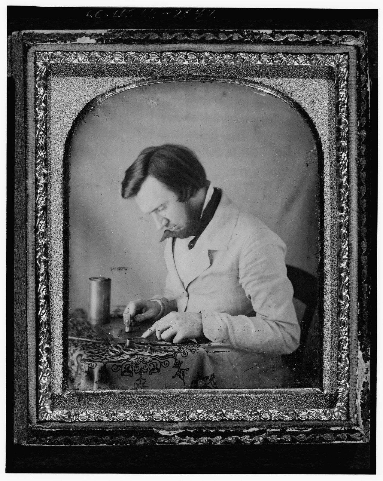 [Occupational portrait of a tinworker (?), half-length, left profile, seated at table with tablecloth, working with a mallet, snips, compass, and metal cylinder], [between 1851 and 1860] — historic photograph