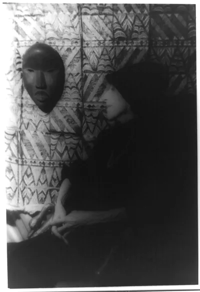 [Portrait of Isak Dinesen (Baroness Karen Blixen) looking at African mask and textile on wall]