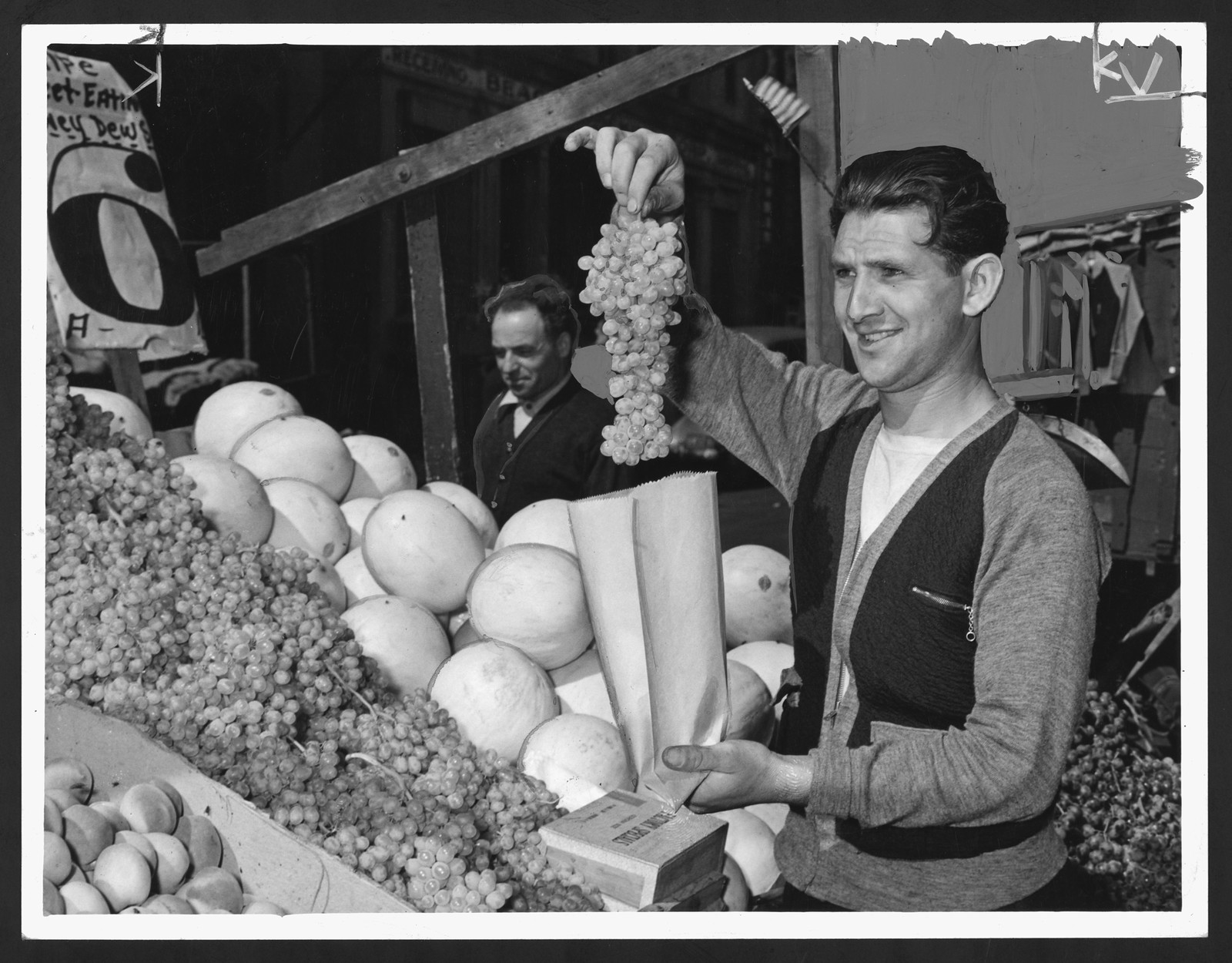 Smiling market vendor holding grapes beside melons and honeydews — historic image