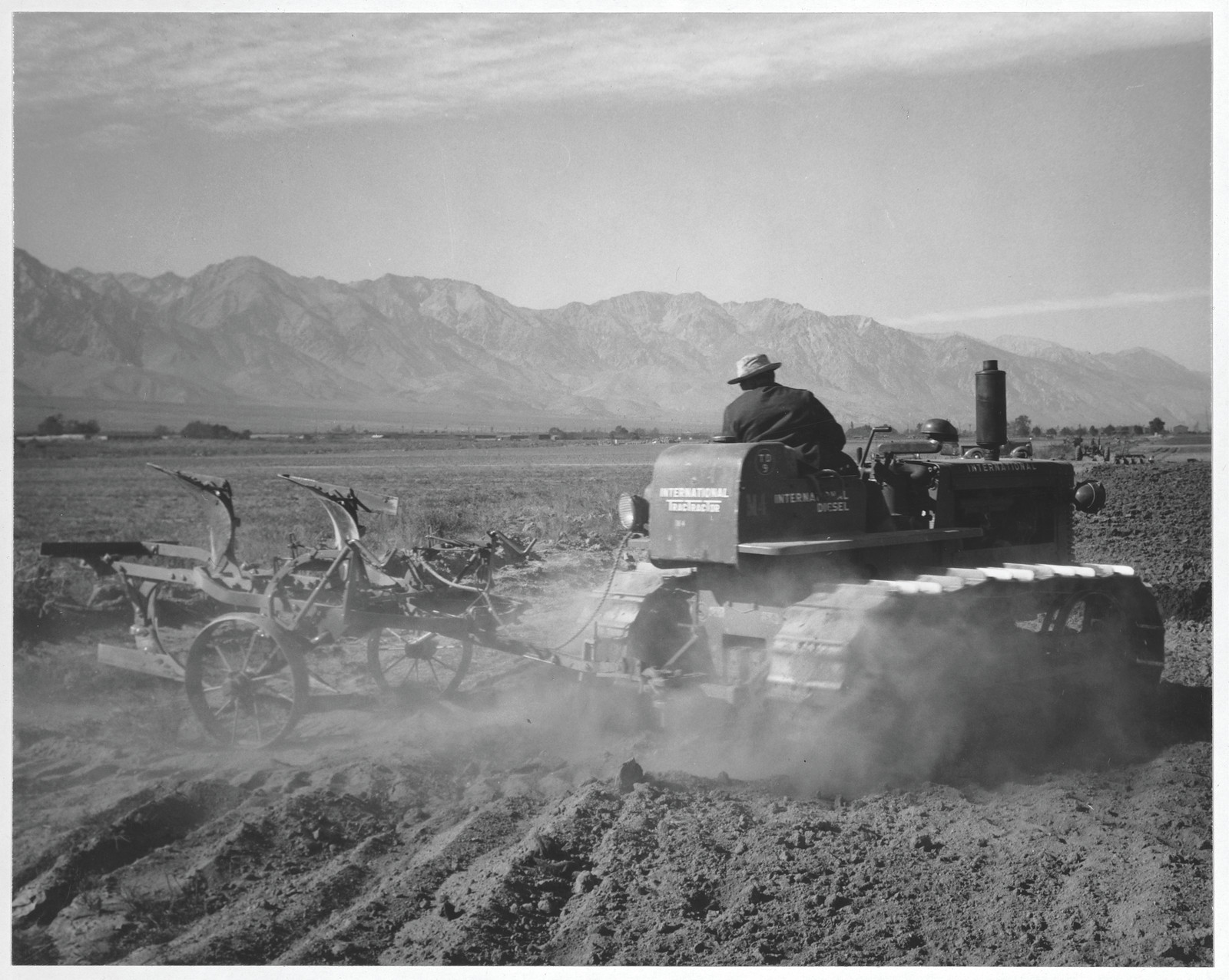 Tractor plowing a field below mountains, black and white photograph — historic photograph