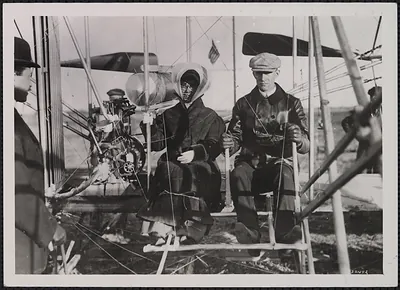 Two people seated in an early biplane, black and white photograph