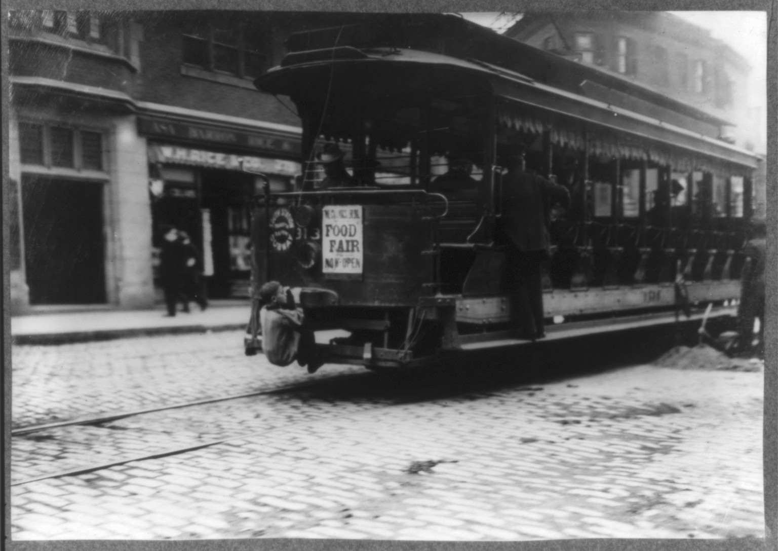Boy clinging to rear of streetcar on cobblestone street — historic image