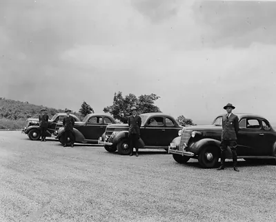 Four uniformed men beside parked 1930s sedans on a gravel overlook
