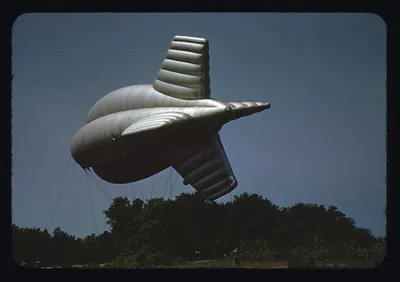 Silver finned barrage balloon above trees under blue sky