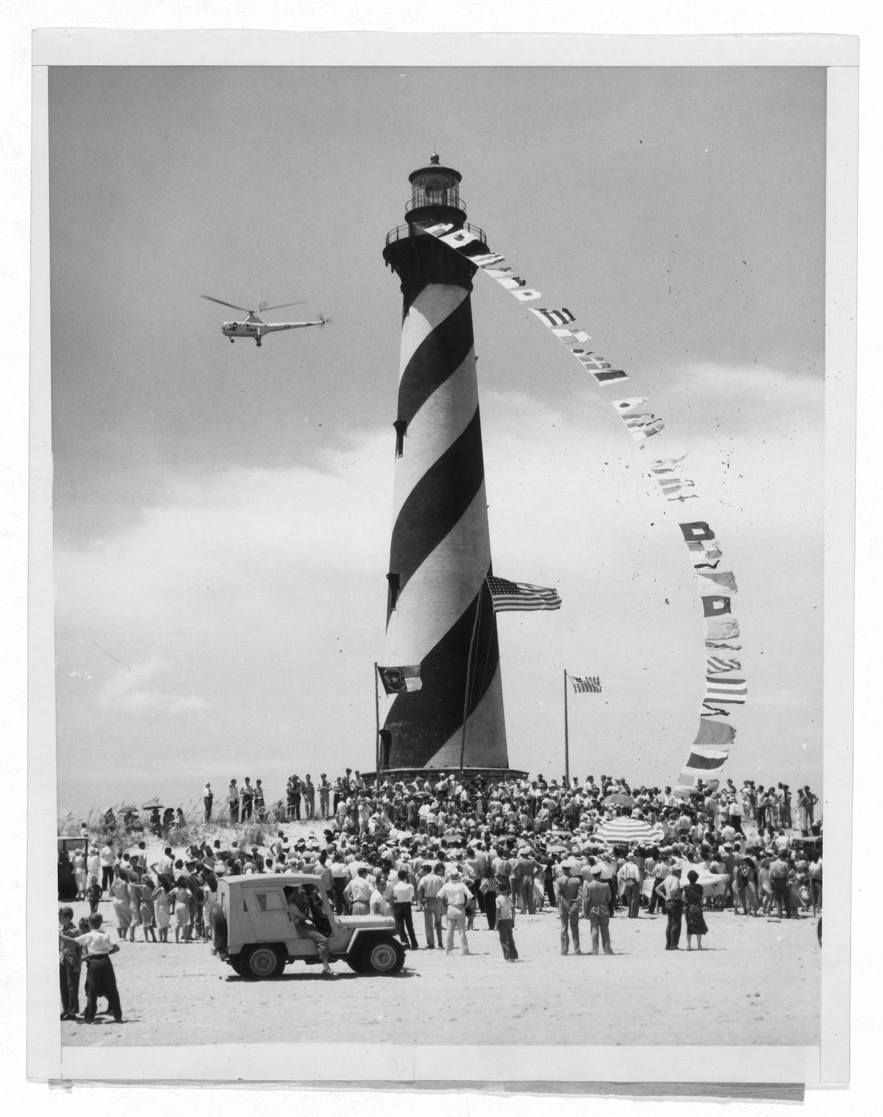 Crowd gathered at striped lighthouse with helicopter overhead — historic image